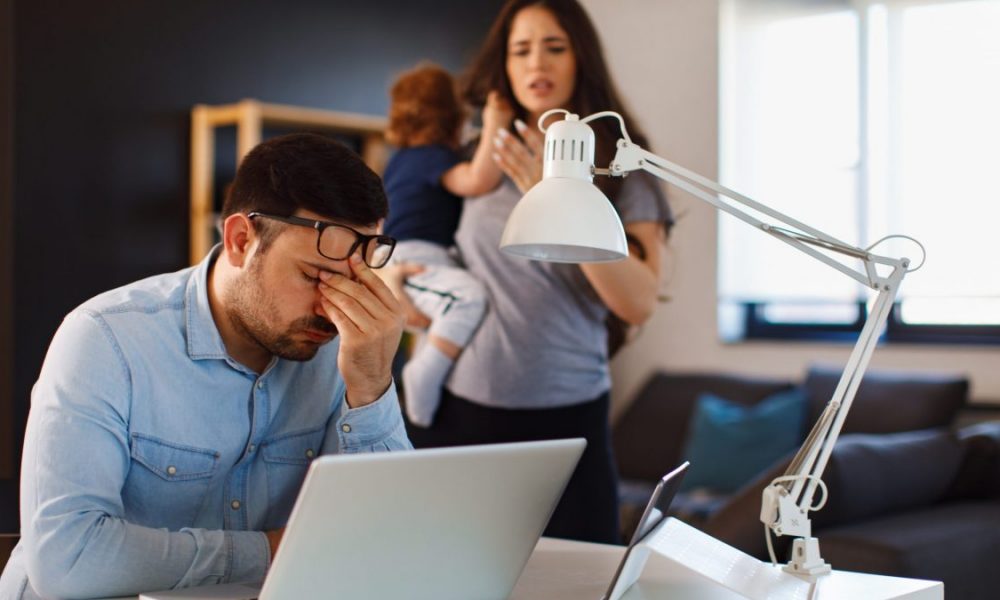 Young family couple having argue about finances at home the husband sits anxious with the laptop and the woman holds the child and complains to him