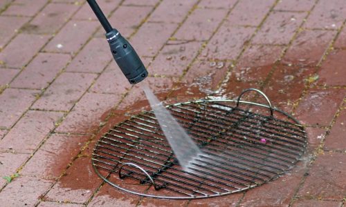 A person uses a pressure washer to clean the grates of an outdoor grill.