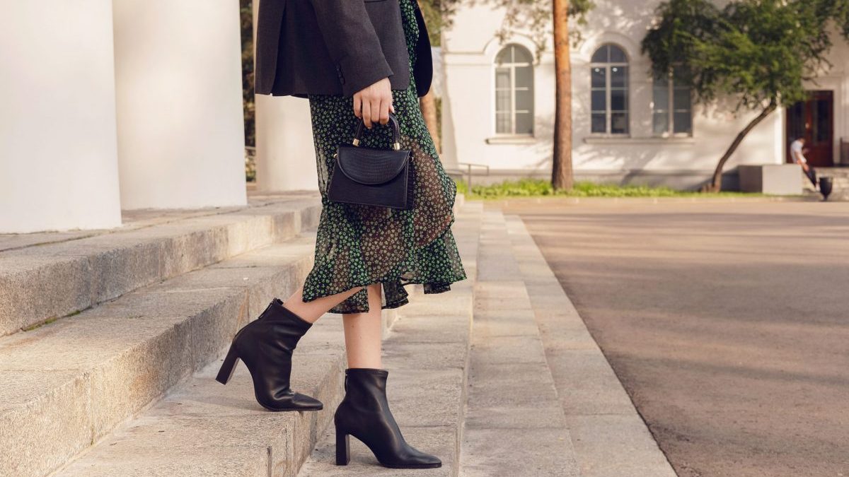 A woman poses in black ankle boots and a long skirt on outdoor stairs.