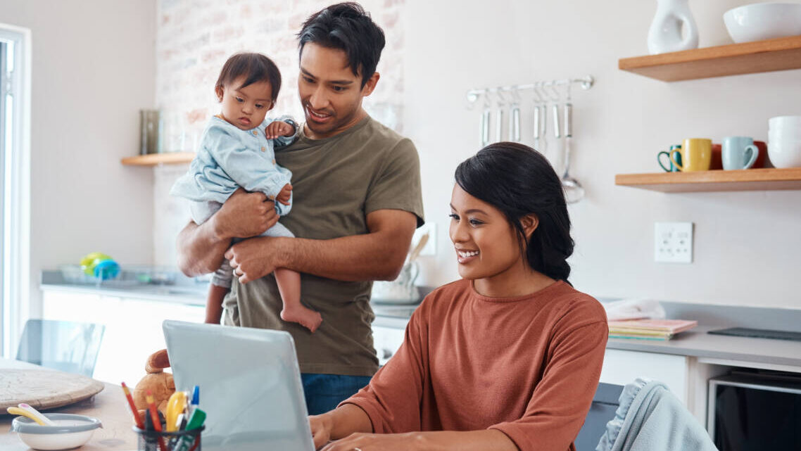 happy parents, dad holds baby while mom works