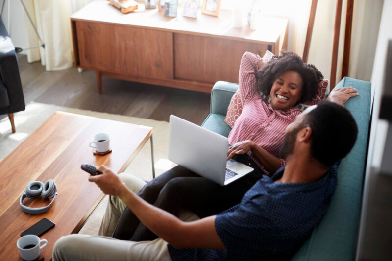 Couple Sitting On Sofa At Home Using Laptop Computer And Watching TV